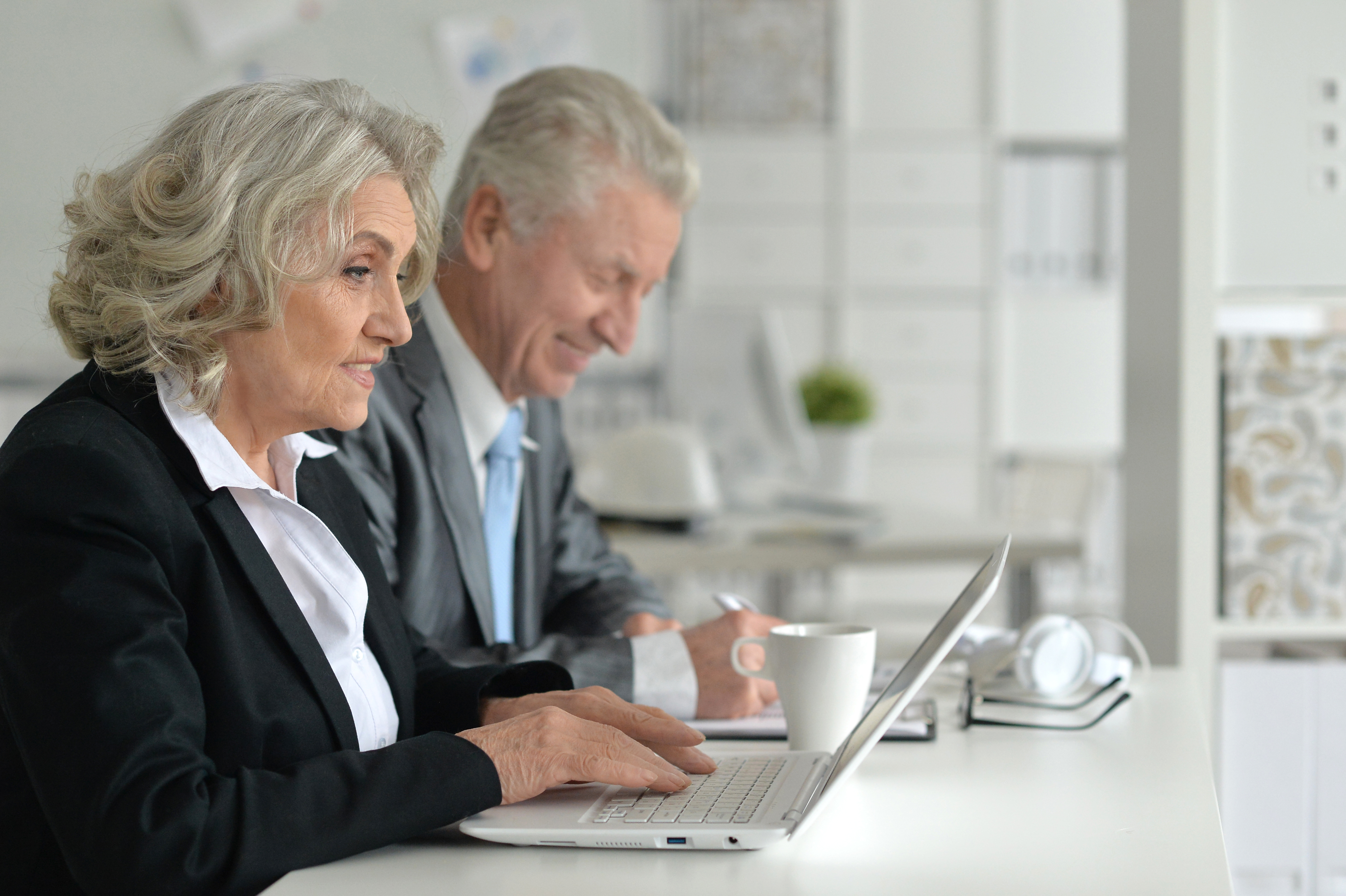 Photo of people looking older, woman and man, white-skinned, lined faces, gray hair, wearing suits, laptop, notetaking on paper.