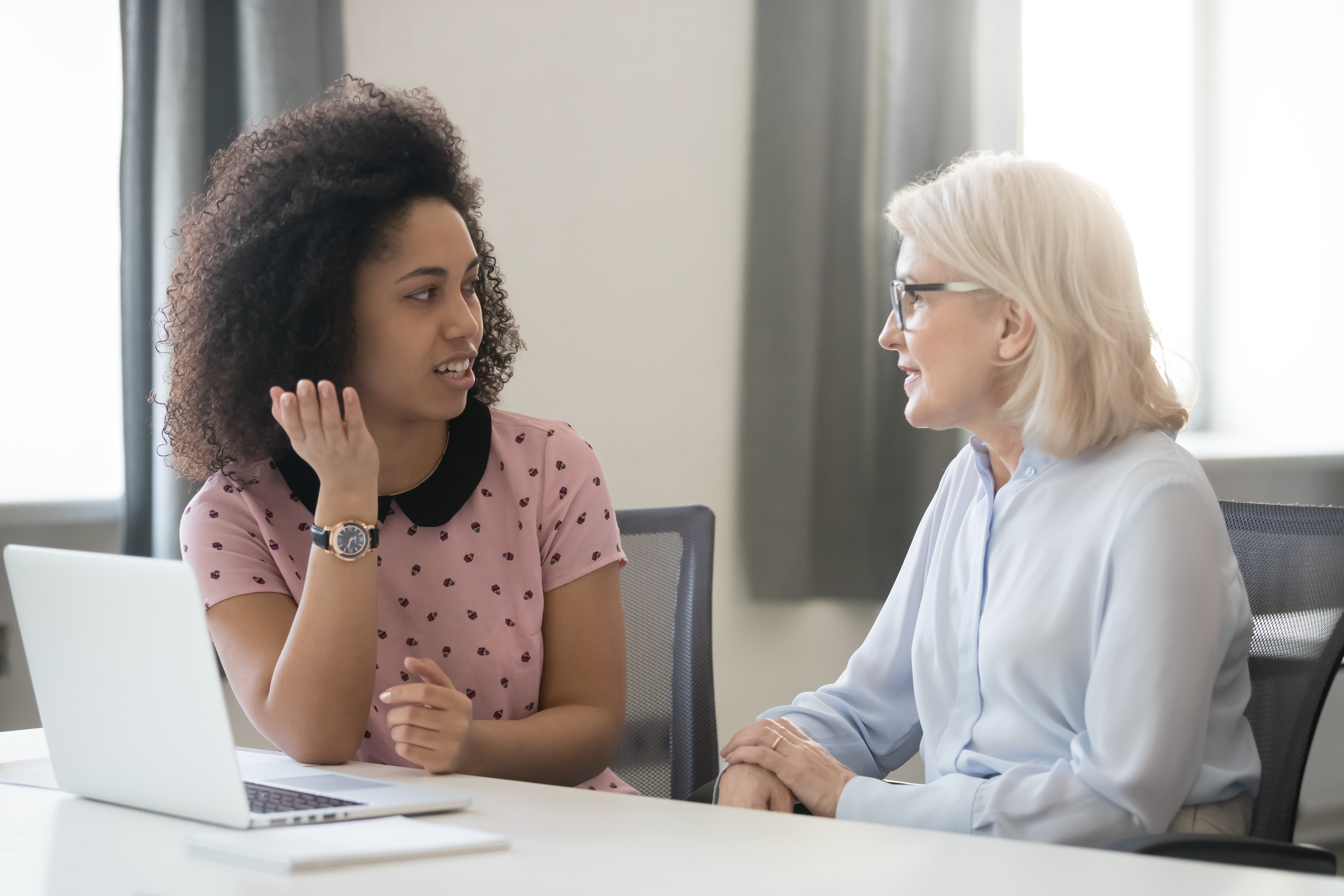 older-appearing woman, white,  with grayish hair talking with younger-appearing woman, black, at table with laptop, office setting