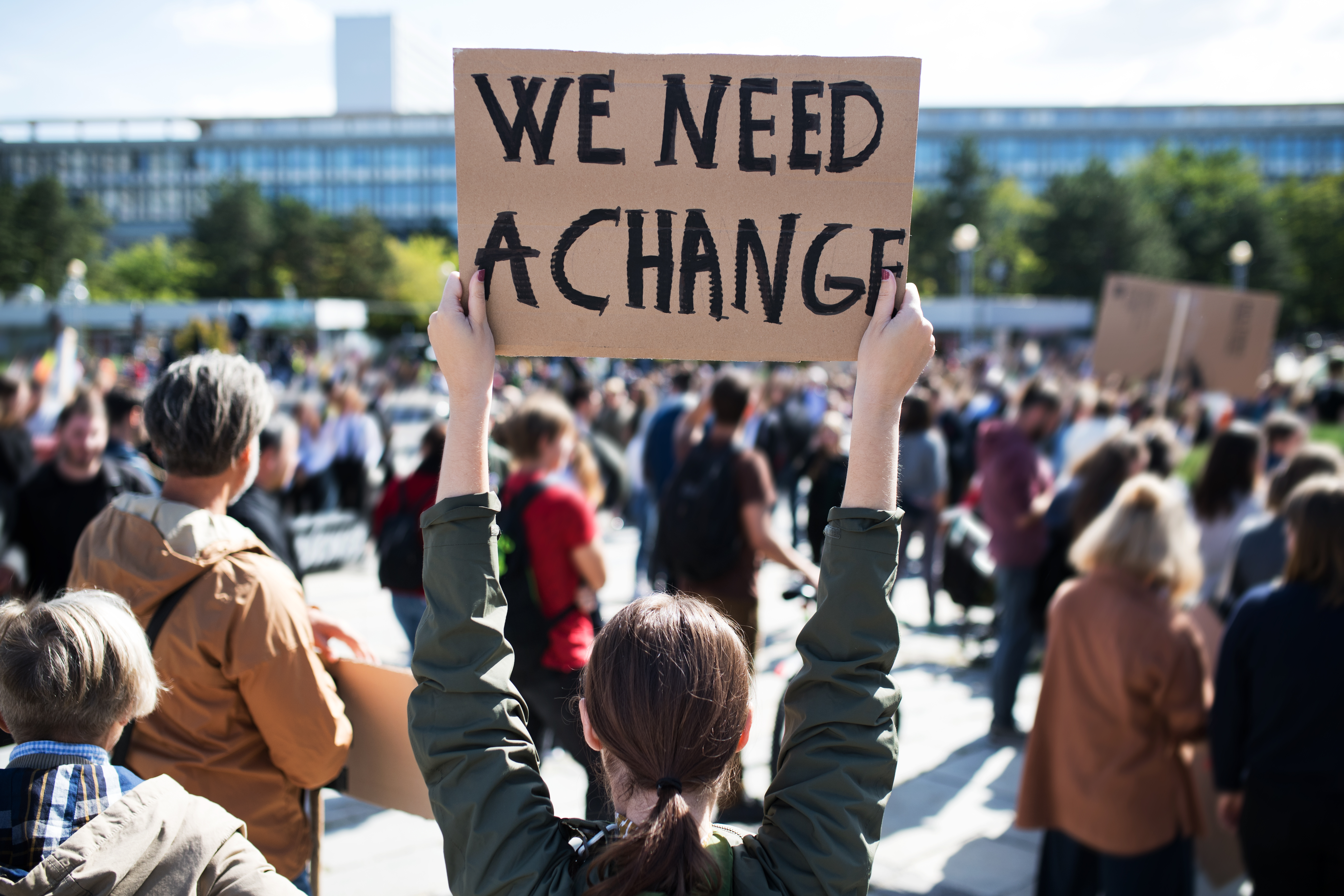 Photo of gathering of people facing buildings, one holding sign, "We Need a Change."