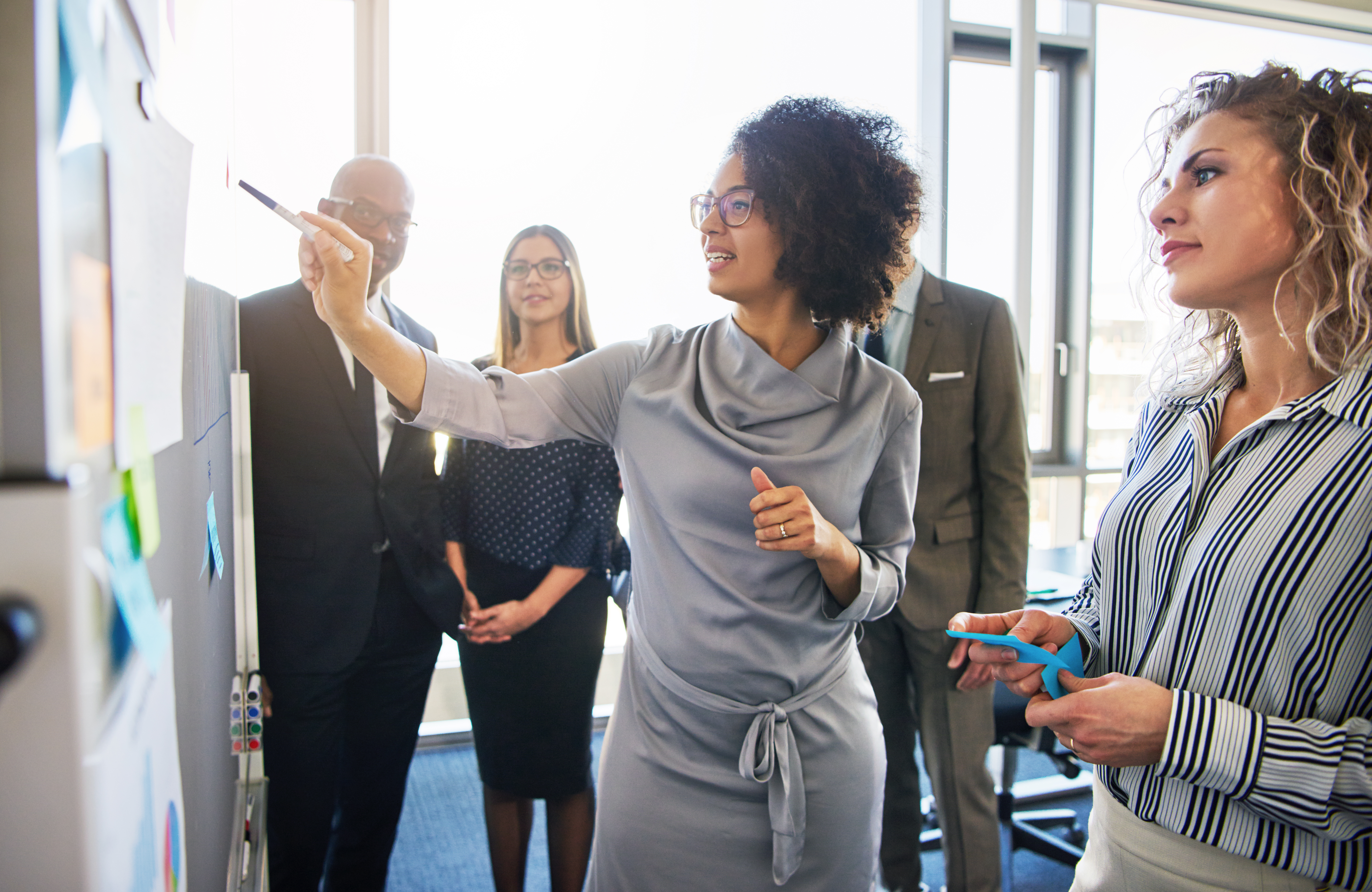 photo appears of woman, black-skin, stylishly, professionally dressed, pointing at chart within group o people listening, mixed color, mostly appearing young