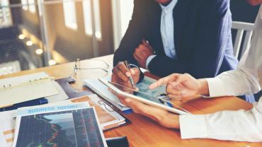 Business people discussing what is on an iPad. Sitting at a table with several files and papers.