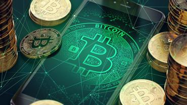 bitcoin logo on a phone screen surrounded by stacks of real bitcoin coins