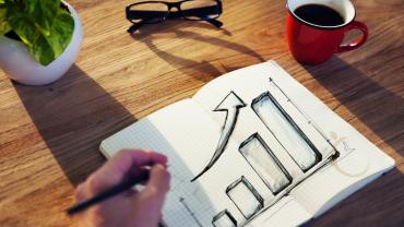 a lefthanded person draws a bar chart and a large arrow trending up in a notebook, there is also a pair of glasses, a small plant, and a cup of coffee on the table