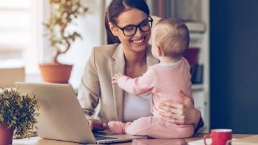 a baby is seated on a desk, held by her smiling mother while simultaneously works on her computer