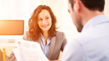 a female professional is facing the viewer with a male's back to the viewer holding papers during an interview