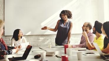 standing woman in front of the white board speaking to her team seated at the table