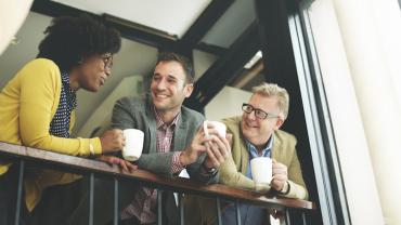 three people lean on a railings holding coffee cups chatting friendlily 