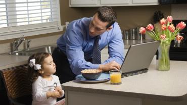 a father in his shirt and tie at the kitchen island leans over his computer looking at his daughter eating breakfast