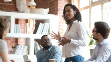 young black woman stands and speaks to a room of diverse colleagues