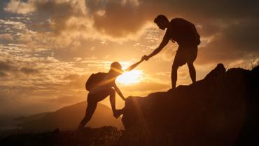 Silhouettes of one hiker helping another hiker up the summit in front of an orange sunset.