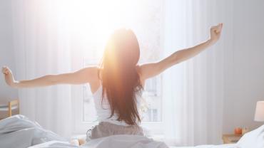 Back view of woman stretching her arms outward in the morning as she wakes up in front of white curtains and the sunlight pouring in.