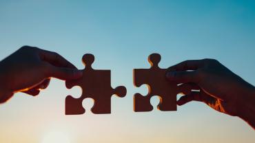 Silhouette of two hands putting two puzzle pieces together in front of a blue and yellow sky at sunset.