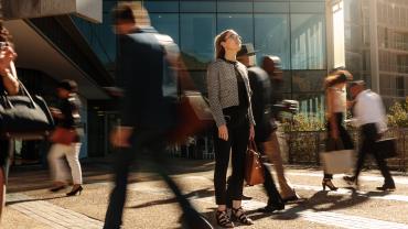 looks to be woman, white, business dress, in front of office-looking building, looking up and in focus compared to people around her