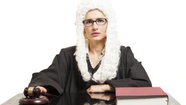 judge with gavel and book, at desk, wearing robe and British-style white wig, appears female/white