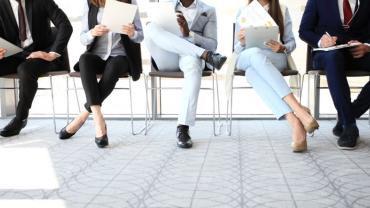 below heads, wearing pants, sitting in chairs, different skin colors, looking at papers, as though in line for job interview