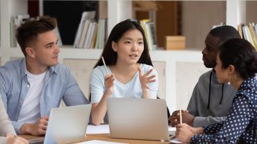 person talking around laptops with three people appearing to be listening intently, mixed genders and skin colors
