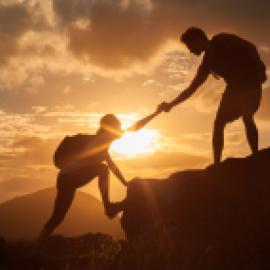 Silhouettes of one hiker helping another hiker up the summit in front of an orange sunset.