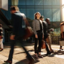 looks to be woman, white, business dress, in front of office-looking building, looking up and in focus compared to people around her