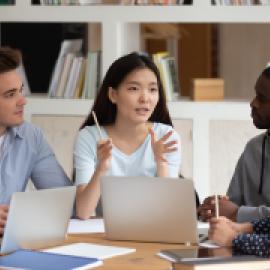 person talking around laptops with three people appearing to be listening intently, mixed genders and skin colors