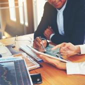 Business people discussing what is on an iPad. Sitting at a table with several files and papers.