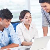 two professionals, a female and male employee sit at a desk looking at a computer while a third employee stands next to them