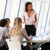 young professional woman standing up at a table with her team seated around her