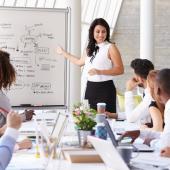 woman standing at a dry erase board giving a presentation to a group seated at the table