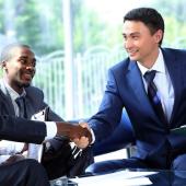 There are five people visible. a woman smiling on the far right. To her left is a man about to stand up shaking hands with another man seated across from him. In between these 2 men are a woman and another seated man all wearing business suits. 