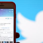 A phone with the twitter profile on screen with the Twitter logo in the background