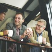 three people lean on a railings holding coffee cups chatting friendlily 