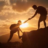 Silhouettes of one hiker helping another hiker up the summit in front of an orange sunset.