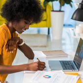 Happy African American business woman smiling and wearing a yellow shirt while sitting at a home office desk in front of a laptop next to white flowers and a yellow couch in the background.