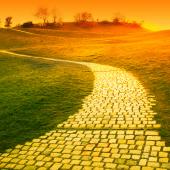 Yellow brick road winding into the distance of a green field toward an orange sunset.