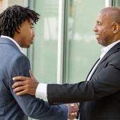 two males in suits, Black, shaking hands, one appearing older has hand on arm of other