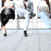 below heads, wearing pants, sitting in chairs, different skin colors, looking at papers, as though in line for job interview