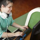 woman, brown or white, it appears, sitting at desk using computer, has headphones and a non-standard keyboard, may be for braille