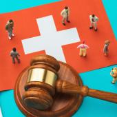 Large Swiss flag with gavel laying across it and small people standing around the gavel. 